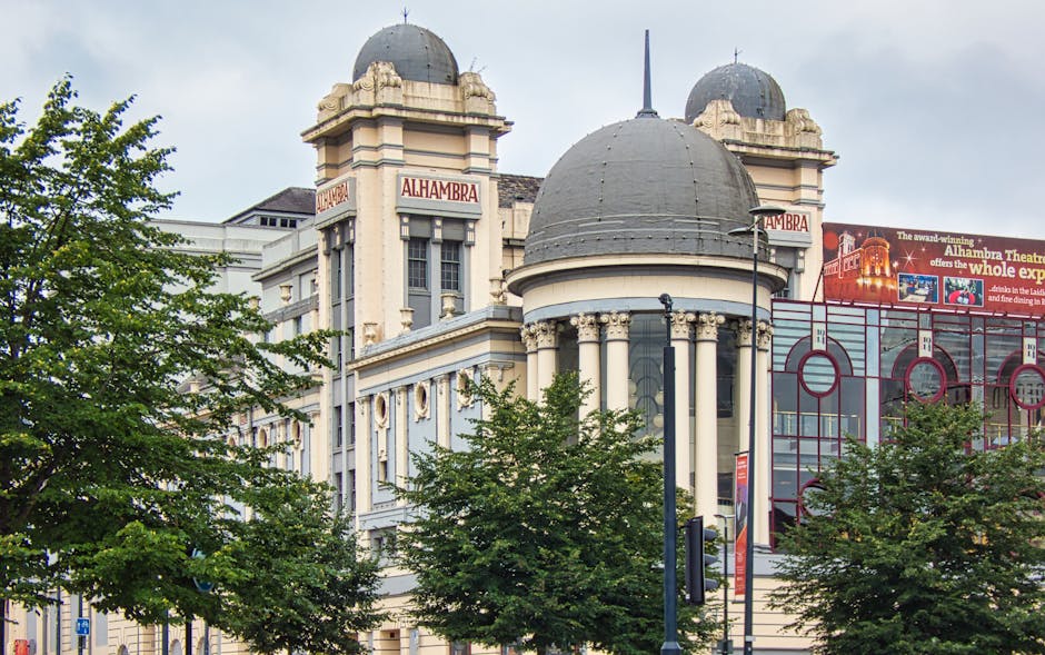 A photograph of the historic Alhambra Theatre building in Haringey, London, showcasing its distinctive architectural features such as tall columns, decorative arches, and a large, rounded dome on top, with two smaller domes on either side. The facade of the building is painted in light colors, with signage displaying 'Alhambra' in prominent red letters. In the foreground, several green trees partially conceal the lower part of the structure, and a black lamppost is visible near the street. To the right of the building, a modern glass-fronted structure with red framing can be seen, along with a large billboard advertising the Alhambra Theatre, highlighting the venue's entertainment offerings. The scene appears to be taken during daylight hours with natural lighting, and the environment suggests an urban setting suitable for home relocation or furniture transport activities, with Movers Haringey providing professional moving services in the area.