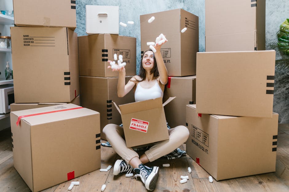 A young woman sitting cross-legged on a wooden floor inside a room during a house move, surrounded by several large cardboard moving boxes, some sealed with red packing tape and one open with a red 'Fragile' sticker attached. She is smiling brightly as she tosses white packing peanuts into the air, which are scattered around her and the boxes. The background includes a textured wall with a blue and grey mottled pattern, a potted green plant, and a small white table with bottles and other items. Visible within the box in her lap is a mix of packing materials, and she is dressed casually in a white tank top, beige pants, and black-and-white sneakers. This scene depicts the packing or loading process typical of a home relocation service, with [COMPANY_NAME] providing professional removals in the Haringey area, as suggested by the context of the property. The image emphasizes the careful handling of household items and packing materials involved in furniture transport and moving logistics.