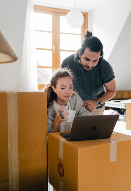A man and a young girl are inside a well-lit room with natural light coming through a large window, which features wooden window frames and a white wall. They are surrounded by cardboard boxes, some sealed with packing tape, indicating an ongoing home relocation process. The girl is seated at a table, holding a white mug and looking at a laptop screen placed on one of the boxes. The man is standing behind her, leaning over slightly, pointing at the laptop, possibly assisting with the packing and moving activities. The scene captures the logistical aspects of furniture transport and packing preparation, with the focus on careful handling of belongings in a home environment. Movers Haringey, a professional removals company, often supports such loading and packing processes, ensuring safe transportation of household items during house removals or relocations.