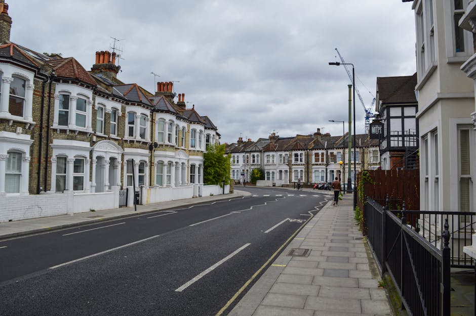 The image shows a quiet residential street in Crouch End N8, with traditional Victorian terraced houses featuring bay windows, brick facades, and tiled roofs under an overcast sky. The pavement runs alongside the row of houses, with a black metal fence visible in the foreground on the right. Street lamps and a few parked cars are present along the curb, and a man is seen walking away from the camera further down the street. The scene is well-lit despite the cloudy weather, with no visible activity related to house removals or loading processes, but the setting reflects a typical area where professional movers like Movers Haringey might operate during home relocations or furniture transport within such neighbourhoods. The overall environment emphasizes the urban residential character suitable for house removals services in the local guide for Crouch End N8.