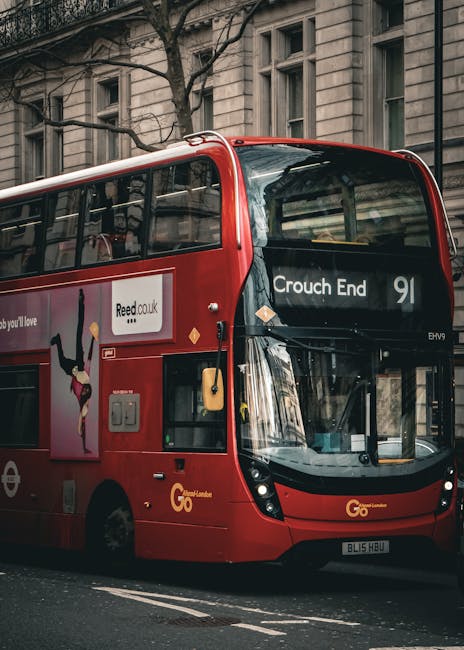 A red double-decker bus parked on a street in front of a historic stone building with multiple windows, indicating a location in Crouch End N8. The bus displays the destination 'Crouch End' on the front electronic sign and has a promotional advertisement on its side panel. Visible inside the bus through the large front windshield and side windows are empty seats and interior lighting. The pavement beside the bus is clear, and the street appears to be in an urban residential area. This scene relates to house removals and moving services by illustrating transportation options used in furniture transport and home relocation processes. Movers Haringey, a professional removals company, might utilize such vehicles for efficient moving logistics, packaging, and loading activities, supporting smooth house move operations in the local area.