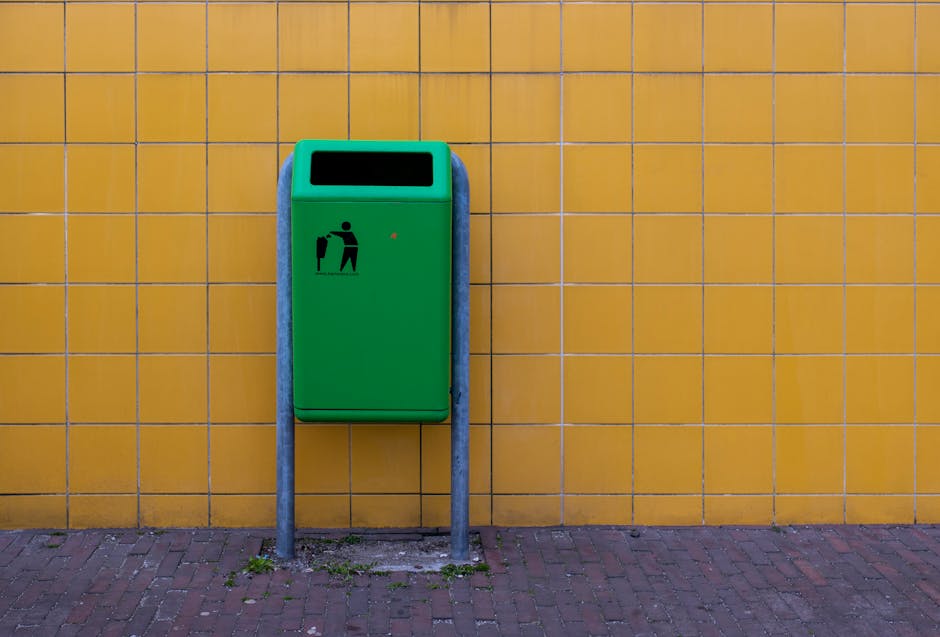 A green public waste bin mounted on two metal posts positioned against a yellow tiled wall outdoors. The bin features a black icon of a person disposing of rubbish into the bin, and has an open rectan