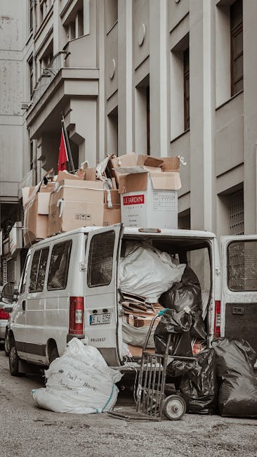 A white commercial van parked on a city street with its rear doors open during a house removal process. The van’s cargo space is filled with various items including large plastic-wrapped furniture, cardboard boxes, and black garbage bags, all arranged for transportation. Several boxes are stacked on the roof of the van, some open and partially crushed, containing packing materials like cardboard and paper. A small hand truck or dolly is positioned next to the open rear doors, loaded with additional boxes and bags. The surrounding environment features tall, light-colored building facades with multiple windows and architectural details, indicating an urban setting. Natural light illuminates the scene, highlighting the loading activity associated with home relocation or furniture transport, with the presence of moving equipment emphasizing the logistical effort by Movers Haringey involved in the packing and loading process, often part of professional removals services related to parking permits for removal vans in Haringey.