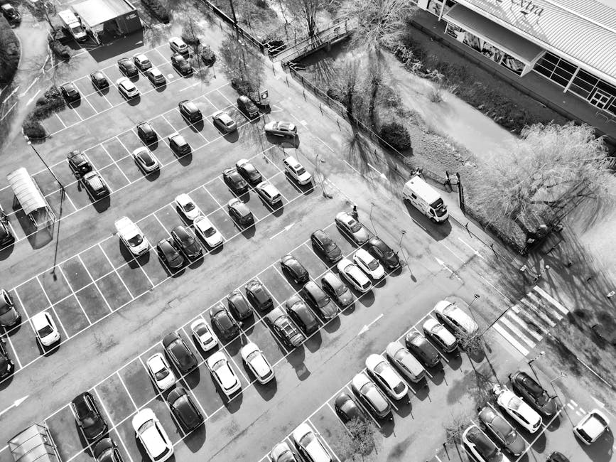 An aerial black-and-white photograph of a parking lot adjacent to a building, featuring multiple rows of parked cars arranged within clearly marked parking spaces. The vehicles are a mix of small to medium-sized cars, with some aligned parallel and others at slight angles. In the background, a white van is seen parked near the corner of the lot, close to a pedestrian crossing marked by white striped lines on the pavement. The parking area is surrounded by a low fence and some trees casting long shadows across the lot, suggesting early morning or late afternoon light. The building on the right has large windows and a modern facade, indicating a commercial or public property. The detailed view captures the organized layout of the parking spaces, with additional areas for potential vehicle movement and passenger access. This image aligns with themes of home relocation and furniture transport, exemplifying the use of parking permits for removal vans, as explained on the Movers Haringey website.