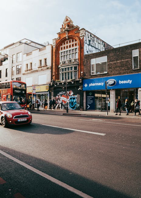 A street scene on Tottenham High Road showing a row of commercial buildings with retail shops and graffiti-covered walls. In the foreground, a red double-decker bus is stopped, while a red car drives