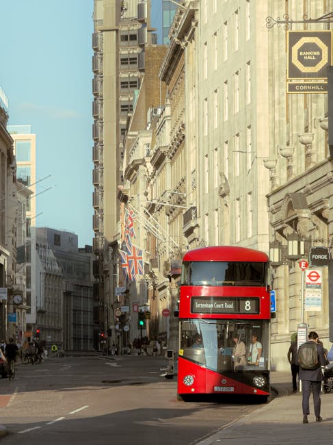 A street scene on Tottenham High Road showing a row of commercial buildings with retail shops and graffiti-covered walls. In the foreground, a red double-decker bus is stopped, while a red car drives