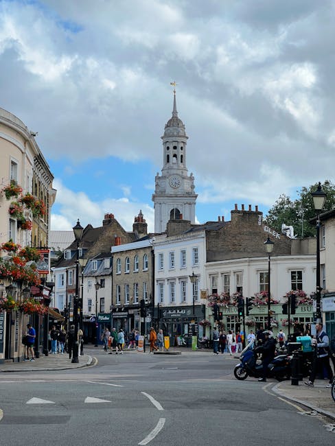 A busy street scene in a historic town centre with multiple older-style terraced buildings featuring retail shops on the ground floors, some with flower baskets hanging from lampposts. Several pedestrians are walking along the pavement, and a few are crossing the road at a pedestrian crossing. In the foreground, vehicles including motorcycles and cars are waiting at the intersection. Behind the buildings, a tall, ornate clock tower with a spire and weather vane at the top rises prominently into a partly cloudy sky. The scene captures the lively atmosphere of a typical urban area suitable for home relocation services, with visible activity and transportation logistics implied by the movement of pedestrians and vehicles. Movers Haringey, a professional removals company, could utilize such an image to illustrate urban moving and furniture transport operations during house removals around the Wood Green N22 area.